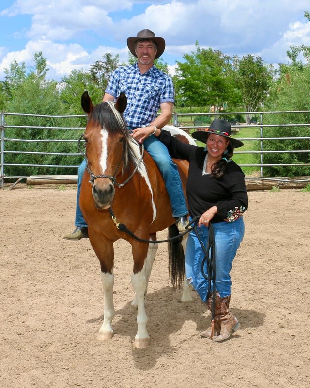 David Kaplan-Gomez smiling in a cowboy hat on a brown and white Paint horse, with Carla standing beside them holding the lead rope in the arena at SonFlower Ranch