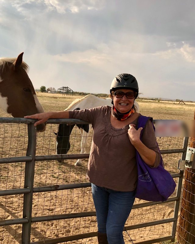 Rose Coca smiling in a riding helmet, leaning on a ranch gate beside a brown and white Paint horse