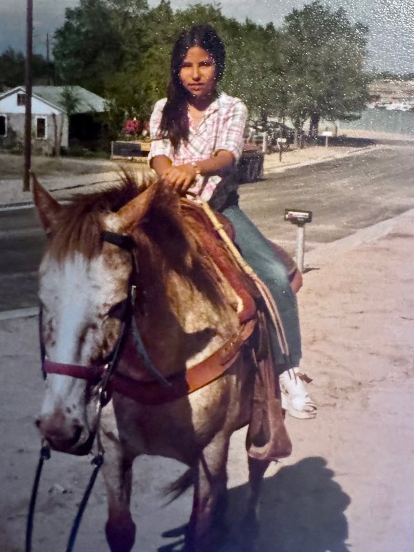 Young Carla in a pink plaid shirt sitting tall on her uncle's brown and white horse on a street in Pueblo, Colorado